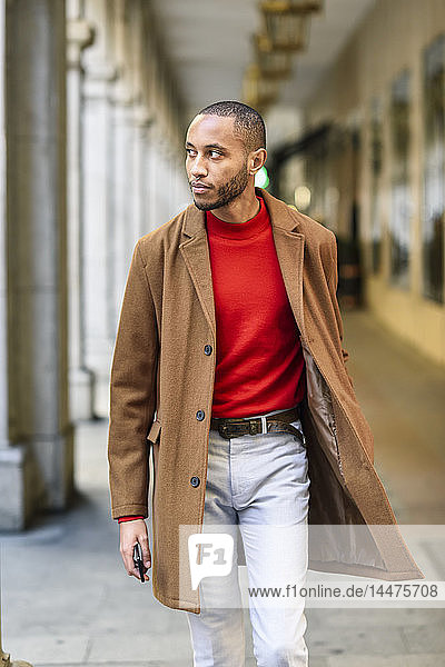 Fashionable young man wearing red pullover and brown coat walking along arcade