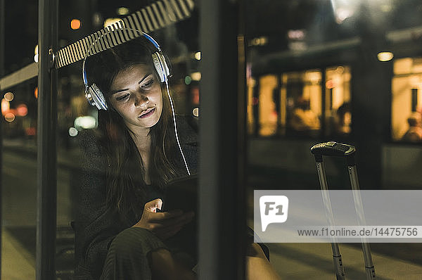 Portrait of young woman with headphones waiting at the station by night using tablet
