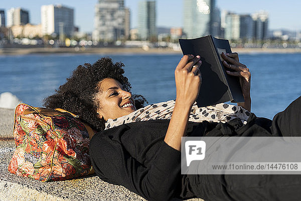 Spanien  Barcelona  lächelnde schwarz gekleidete Frau entspannt sich auf einer Mauer vor dem Meer