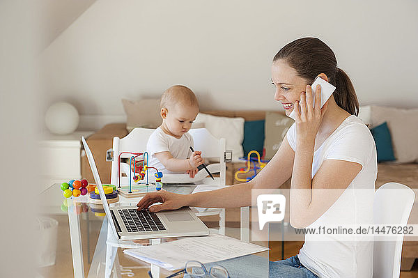 Lächelnde Mutter benutzt Laptop und Handy  während die kleine Tochter zu Hause am Tisch spielt