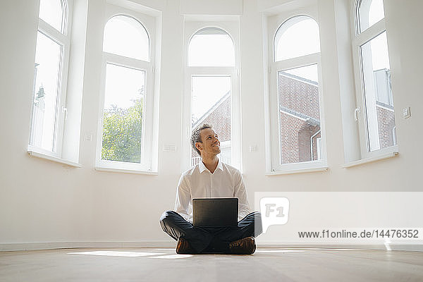 Geschäftsmann sitzt auf dem Boden seines frisch renovierten Hauses  benutzt Laptop und lächelt