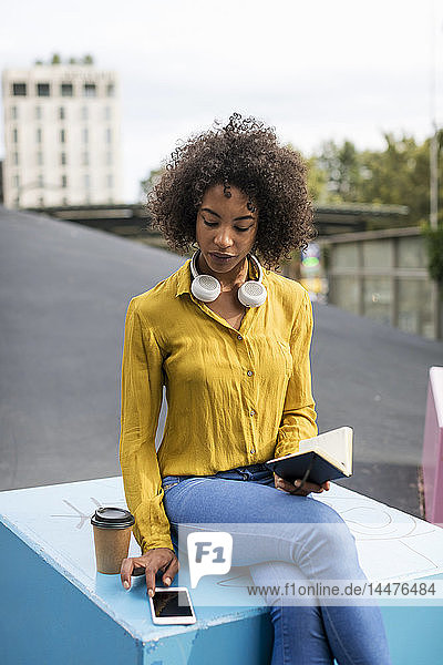 Frau sitzt auf Bank mit Kaffee zum Mitnehmen und Notebook und schaut auf Smartphone