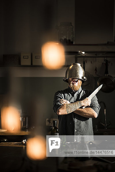 Mann mit Küchenmesser in der Küche stehend  Sieb als Helm tragend