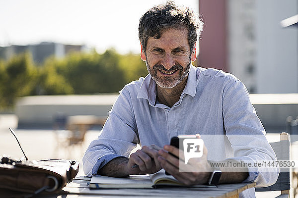 Porträt eines lächelnden reifen Mannes  der mit Handy und Notebook an einem Tisch im Freien sitzt