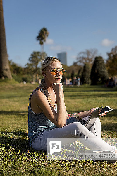 Junge Frau sitzt im Park mit Handy und Laptop
