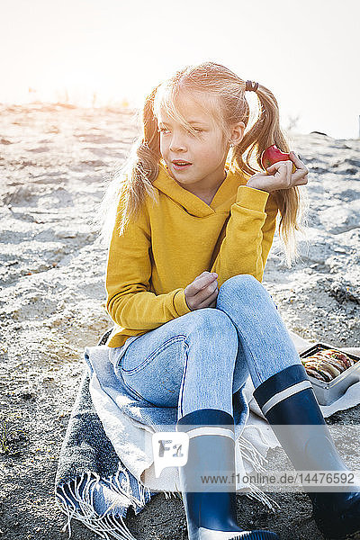 Portrait of girl with plaits sitting on the beach in autumn eating an apple