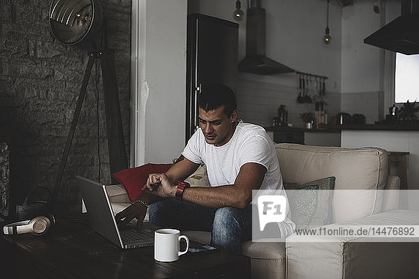 Junger Mann sitzt zu Hause auf dem Sofa  benutzt Laptop und überprüft die Uhrzeit