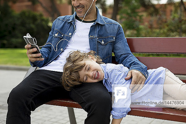 Happy father with son on a bench using cell phone and earbuds