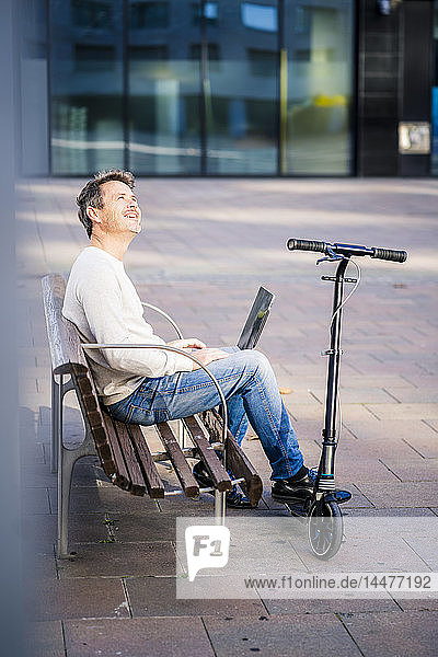 Casual businessman with kick scooter  sitting on a bench  working relaxed in the city