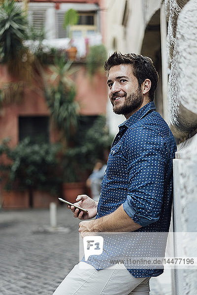 Porträt eines lächelnden Mannes mit Smartphone  der sich an die Fassade lehnt und in die Ferne schaut