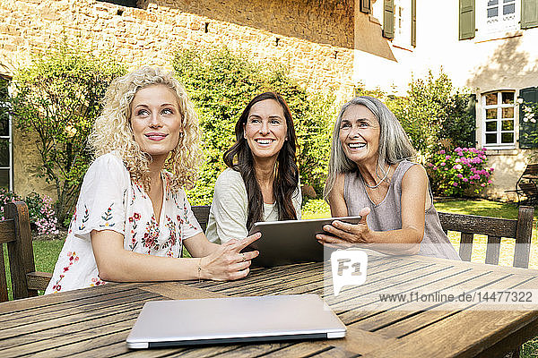 Drei Frauen unterschiedlichen Alters sitzen mit Tabletten am Gartentisch