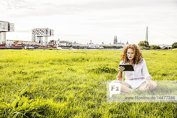 Deutschland  Köln  junge Frau sitzt auf der Wiese und schaut auf die Tablette