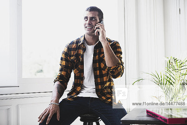 Lächelnder junger Mann  der am Fenster mit dem Handy telefoniert