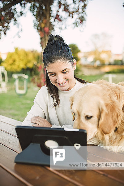 Lächelnde junge Frau mit einer Tablette in einem Park mit ihrem Hund