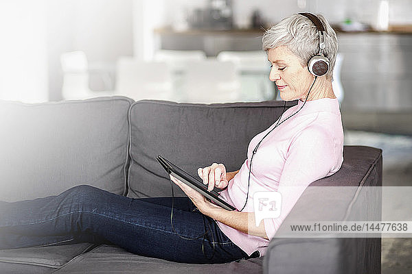 Reife Frau sitzt auf dem Sofa und benutzt ein digitales Tablet  während sie mit Kopfhörern Musik hört