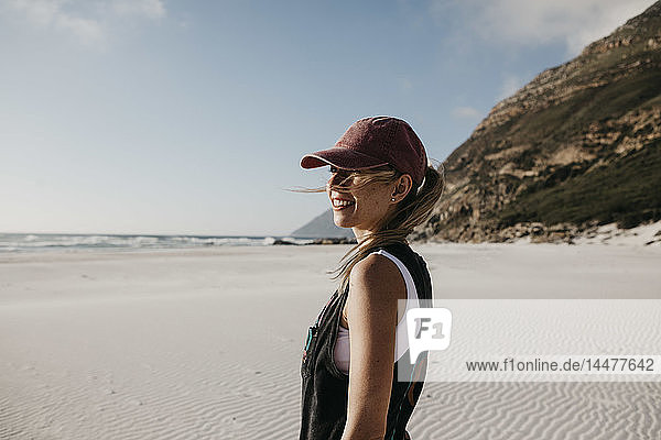 Südafrika  Western Cape  Noordhoek Beach  lächelnde junge Frau mit Basecap am Strand stehend