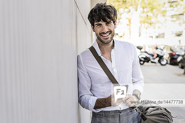 Porträt eines lächelnden jungen Mannes mit Tasche und Handy in der Stadt