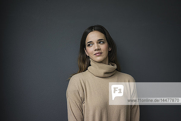 Portrait of smiling woman wearing light brown turtleneck pullover leaning against grey wall