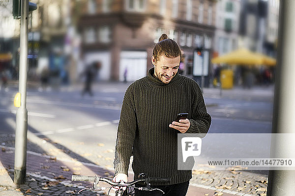 Smiling young man with bicycle in the city looking at cell phone