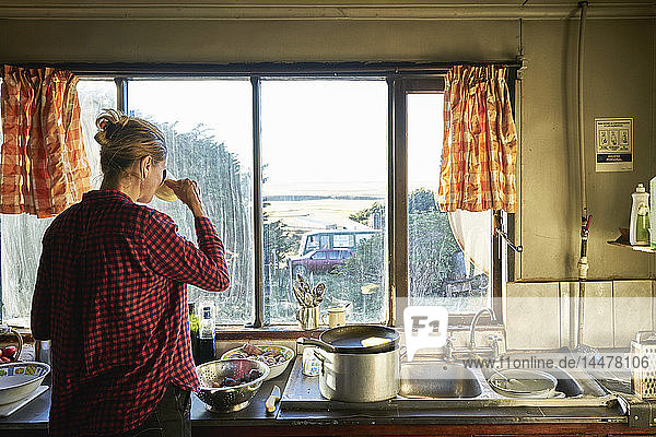 Frau steht in Küche  bereitet Essen vor und trinkt aus Tasse