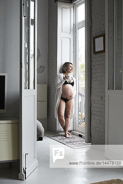 Hübsche blonde schwangere Frau steht am Fenster und hält eine Tasse Tee