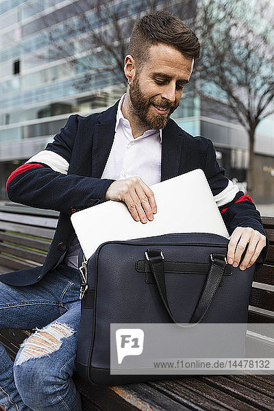 Stylish businessman sitting on bench in the city taking out laptop from bag