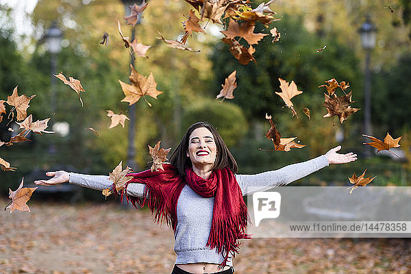 Porträt einer lachenden jungen Frau mit rotem Schal  die Herbstblätter in die Luft wirft