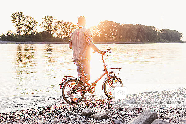 Young man walking with bicycle at the riverbank at sunset