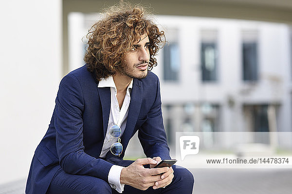 Porträt eines jungen Geschäftsmannes mit lockigem Haar im blauen Anzug auf einer Bank im Freien sitzend