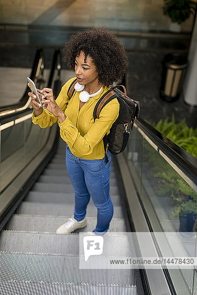 Frau mit Rucksack und Kopfhörern steht mit einem Smartphone auf der Rolltreppe