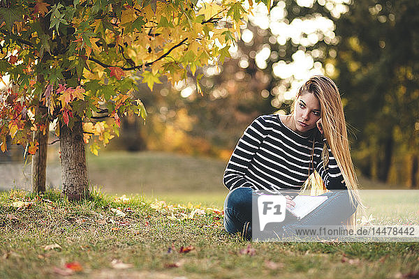 Porträt einer nachdenklichen jungen Frau mit Notizbuch  die im Herbst auf einer Wiese in einem Park sitzt und Notizen macht