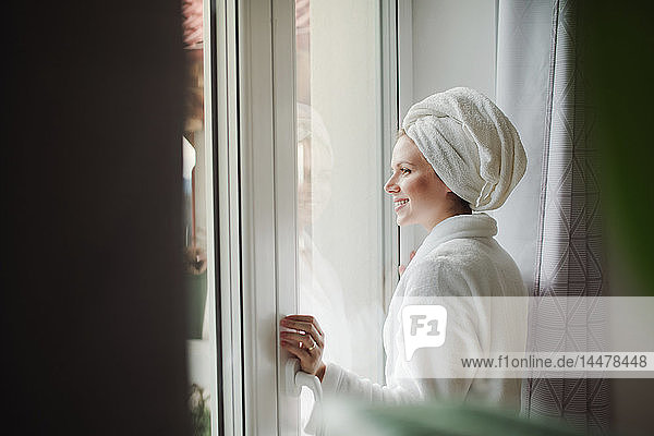 Lächelnde Frau im Bademantel mit Handtuch um den Kopf  die zu Hause aus dem Fenster schaut