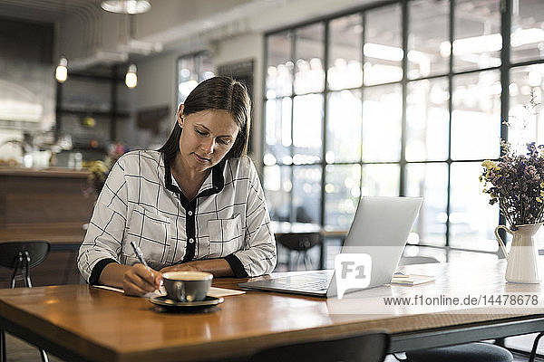 Young businesswoman in a cafe writing on paper and working with laptop on wooden table