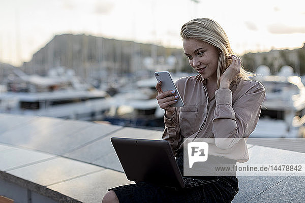 Beautiful young businesswoman using cell phone and laptop at the waterfront