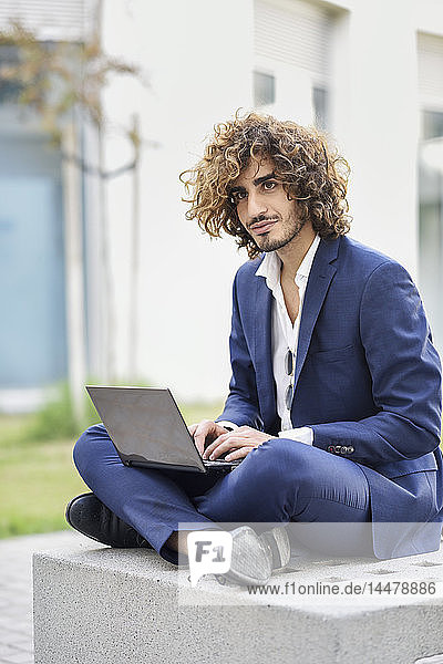 Porträt eines jungen Geschäftsmannes mit lockigem Haar im blauen Anzug  der mit seinem Laptop auf einer Bank im Freien sitzt