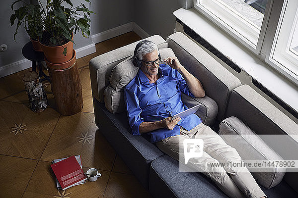 Smiling mature man with tablet and headphones relaxing on couch at home