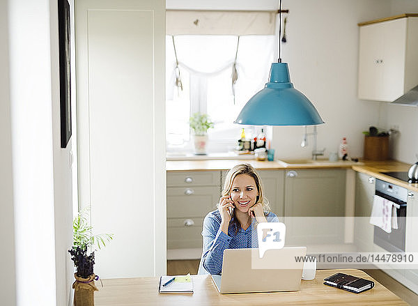 Lächelnde Frau mit Laptop und Handy bei der Arbeit zu Hause