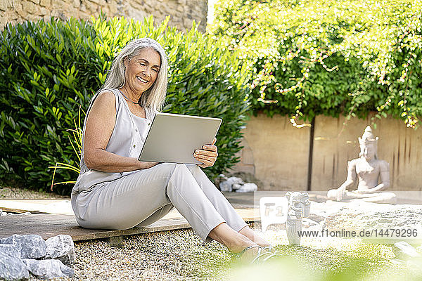 Lächelnde Frau mit langen grauen Haaren sitzt mit Tablette auf Terrasse im Garten