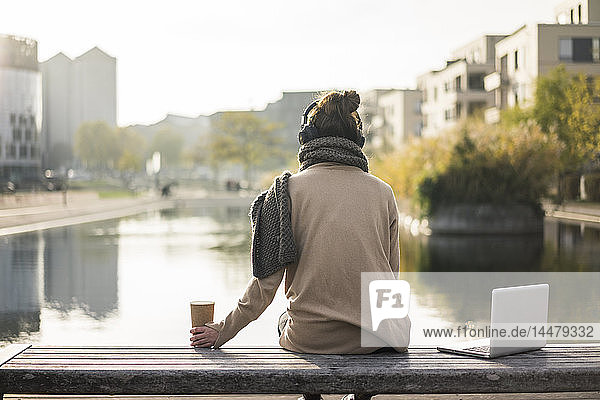 Rückenansicht einer Frau  die auf einer Bank mit Laptop und Kaffee sitzt  um mit Kopfhörern Musik zu hören