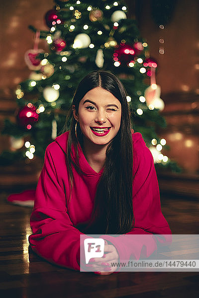 Porträt eines zwinkernden Teenagers  der auf dem Boden vor dem Weihnachtsbaum liegt