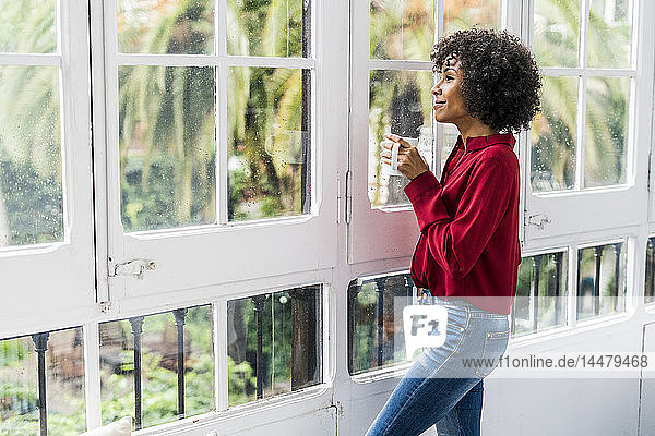 Lächelnde Frau mit Tasse Kaffee  die zu Hause aus dem Fenster schaut