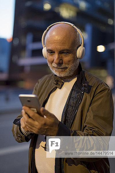 Spain  Barcelona  senior man with headphones and cell phone in the city at dusk