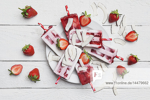 Hausgemachte Erdbeer-Kokos-Eis-Lollies mit frischen Erdbeeren und Kokosnussscheiben auf dem Teller