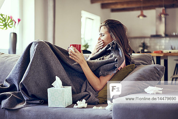Portrait of woman lying sick on sofa coughing and trying to drink hot tea