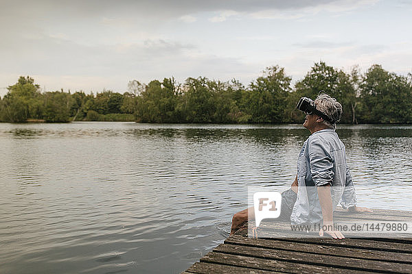 Senior man sitting on jetty at a lake wearing VR glasses