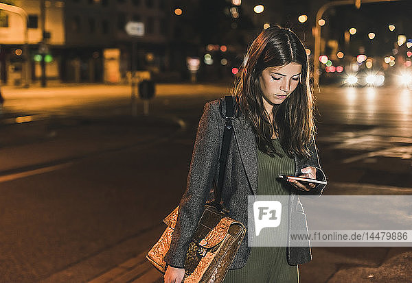 Junge Geschäftsfrau mit Ledertasche betrachtet Handy bei Nacht