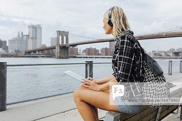 USA  New York City  Brooklyn  junge Frau sitzt am Wasser mit Rucksack  Kopfhörer und Tablett