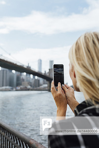 USA  New York City  Brooklyn  junge Frau am Wasser beim Fotografieren der Brooklyn Bridge mit dem Handy