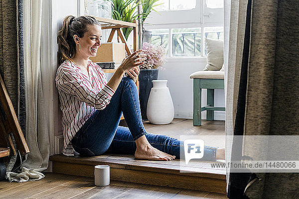 Lächelnde Frau sitzt zu Hause mit Handy auf dem Boden
