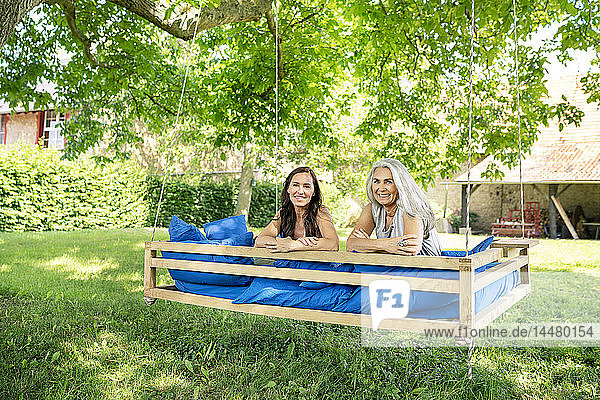 Porträt von zwei lächelnden Frauen  die sich auf einem hängenden Bett im Garten entspannen